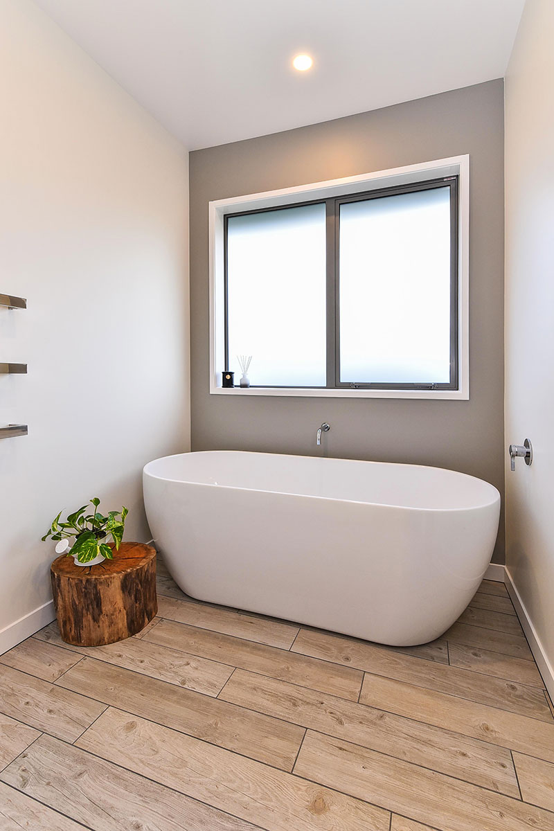 Large Free standing bath in front of grey feature wall with wooden floor tiles, large stump and plant