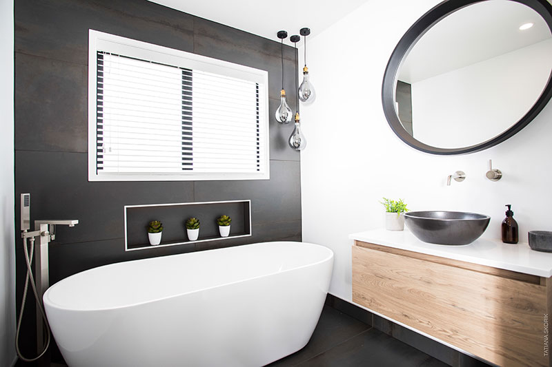 Showhome bathroom with freestanding bath, wooden vanity, feature pendent lights, charcoal basin and charcoal round mirror