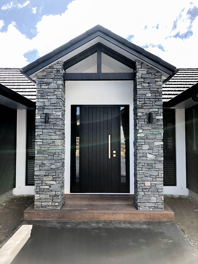Entrance to new home with schist pillars, wooden deck, black front door, white plaster and black weatherboards
