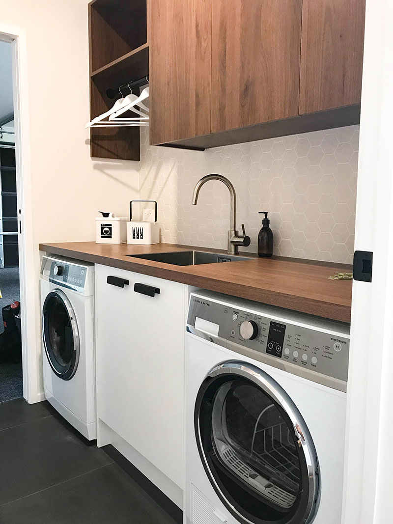Gorgeous timber cabinets & benchtop in showhome laundry at Paerata Rise. Dark tiled floor and offwhite mosaic splashback